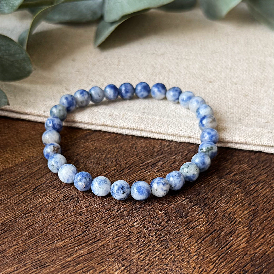 Close-up of the Blue Spot Jasper Serenity Bracelet resting on textured wood and natural linen. The elastic bracelet features uniform polished beads with striking variations of vibrant purple marbling against a creamy white background. Soft-focus eucalyptus creates a serene backdrop, highlighting the bracelet's purpose as a portable oasis of calm. Each unique stone pattern serves as a reminder to maintain balance amid life's challenges.