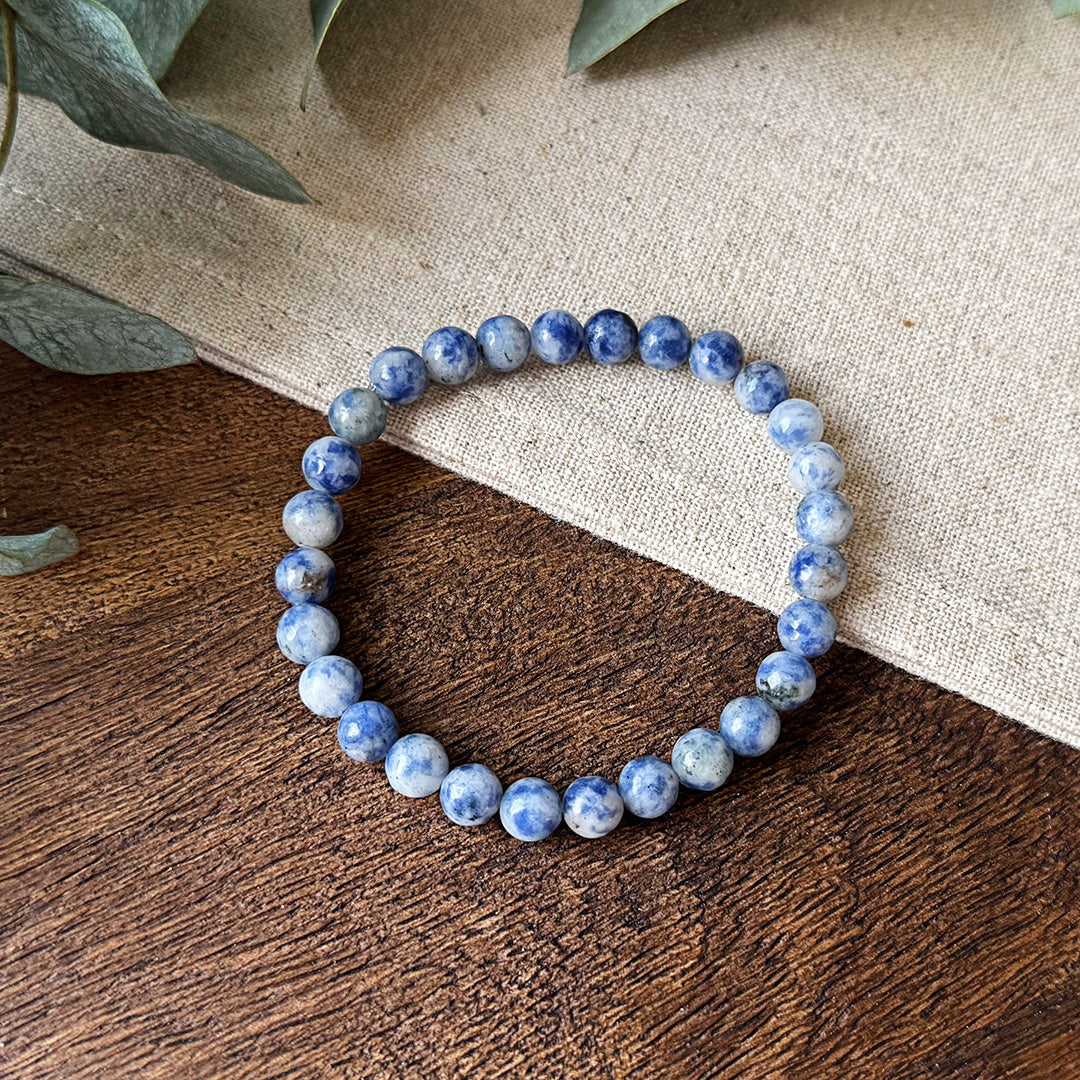 Blue Spot Jasper Serenity Bracelet artfully displayed on a textured wooden surface and natural linen fabric. The polished gemstone beads showcase stunning purple-blue marbling against a white background, with each bead uniquely patterned like tiny galaxies. Fresh eucalyptus leaves frame the corner, enhancing the bracelet's earthy, calming aesthetic. Perfect for those seeking to transform chaos into tranquility while adding a touch of natural elegance to their style.