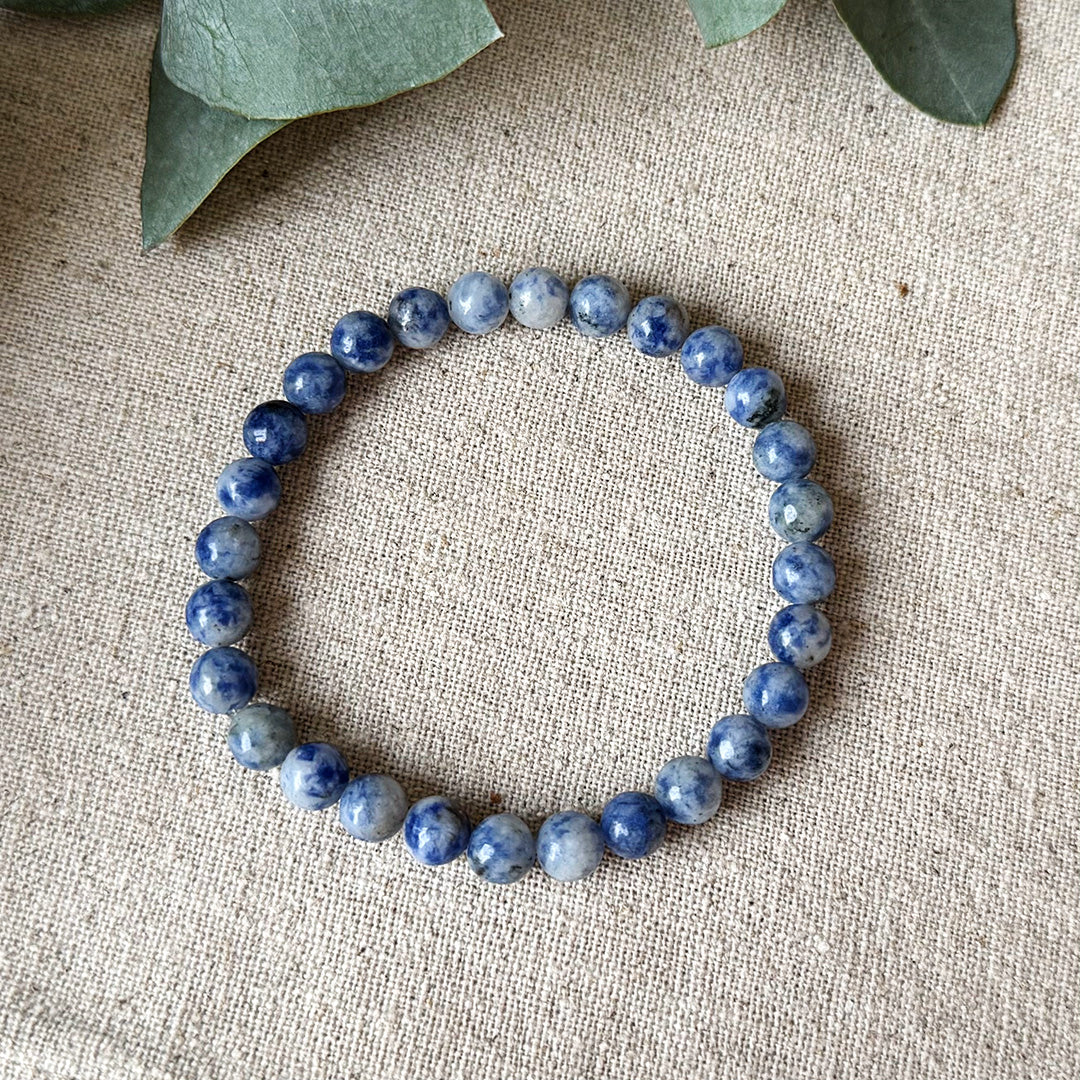 Blue Spot Jasper Serenity Bracelet arranged in a perfect circle on natural linen fabric. Each bead displays mesmerizing swirls of deep purple and soft lavender against white stone, creating a hypnotic pattern that captures the stone's unique energy properties. Subtle eucalyptus leaves peek into frame, complementing the bracelet's soothing nature and emphasizing its connection to calm and balance. An everyday wearable talisman for courage, communication, and inner peace.
