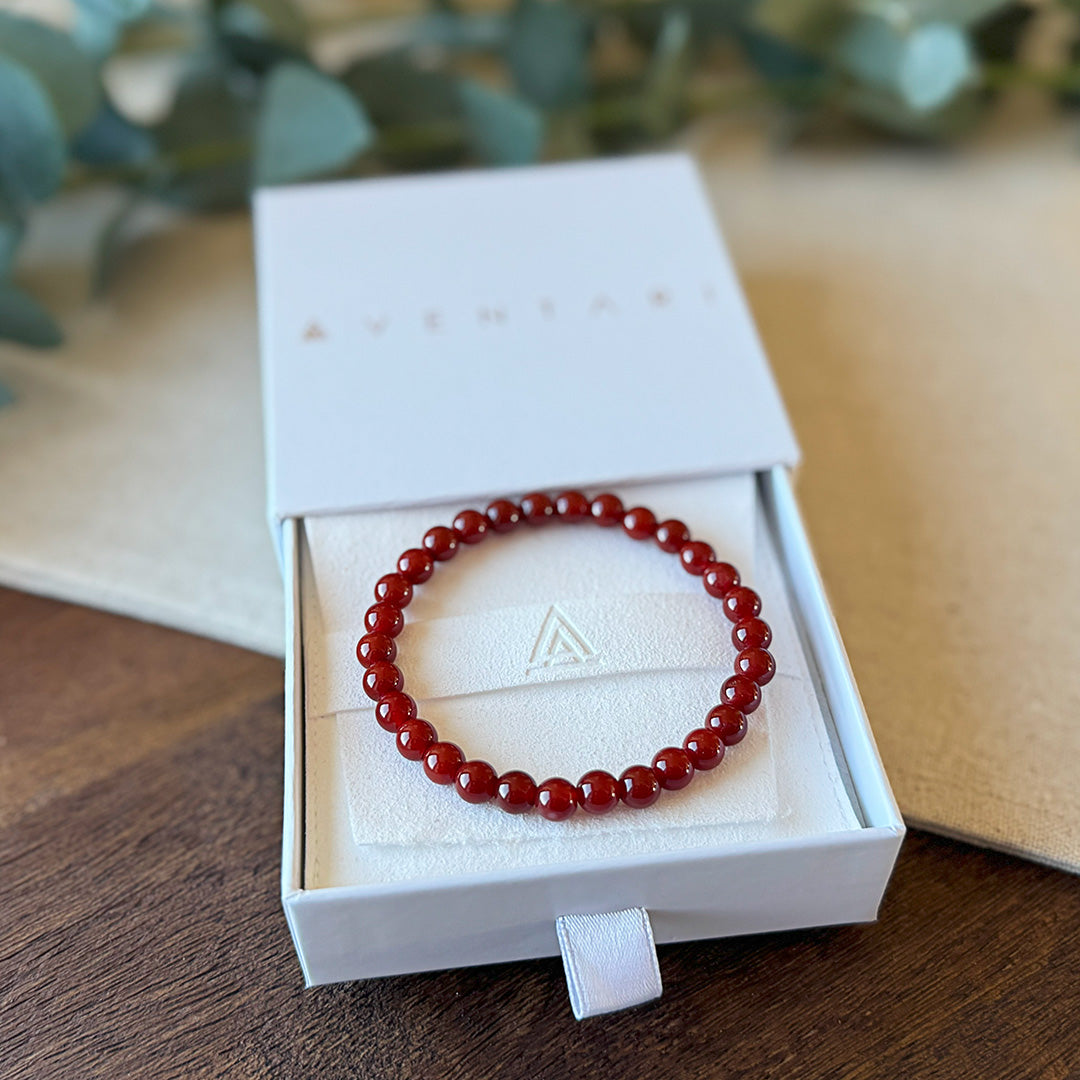 Red Agate Courage Bracelet elegantly presented in an Aventari branded gift box. The sleek white packaging with gold lettering displays the bracelet arranged on a cream-colored cushion embossed with the brand's triangular logo. The vibrant crimson beads form a perfect circle, showcasing their consistent size and exceptional polish. Eucalyptus leaves create a natural backdrop, emphasizing the bracelet's connection to earth energy while adding a touch of sophistication to the presentation. An ideal gift for in