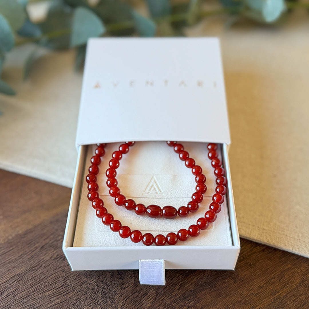 Red Agate Courage Necklace presented in a premium Aventari branded gift box. The elegant white packaging with gold lettering reveals the necklace beautifully arranged on a cream-colored cushion embossed with the brand's triangular logo. The vibrant red gemstone beads catch the light with their glossy finish, highlighting the stone's fiery energy and protective qualities. An ideal gift for someone who needs a boost of confidence and courage to face life's challenges.
