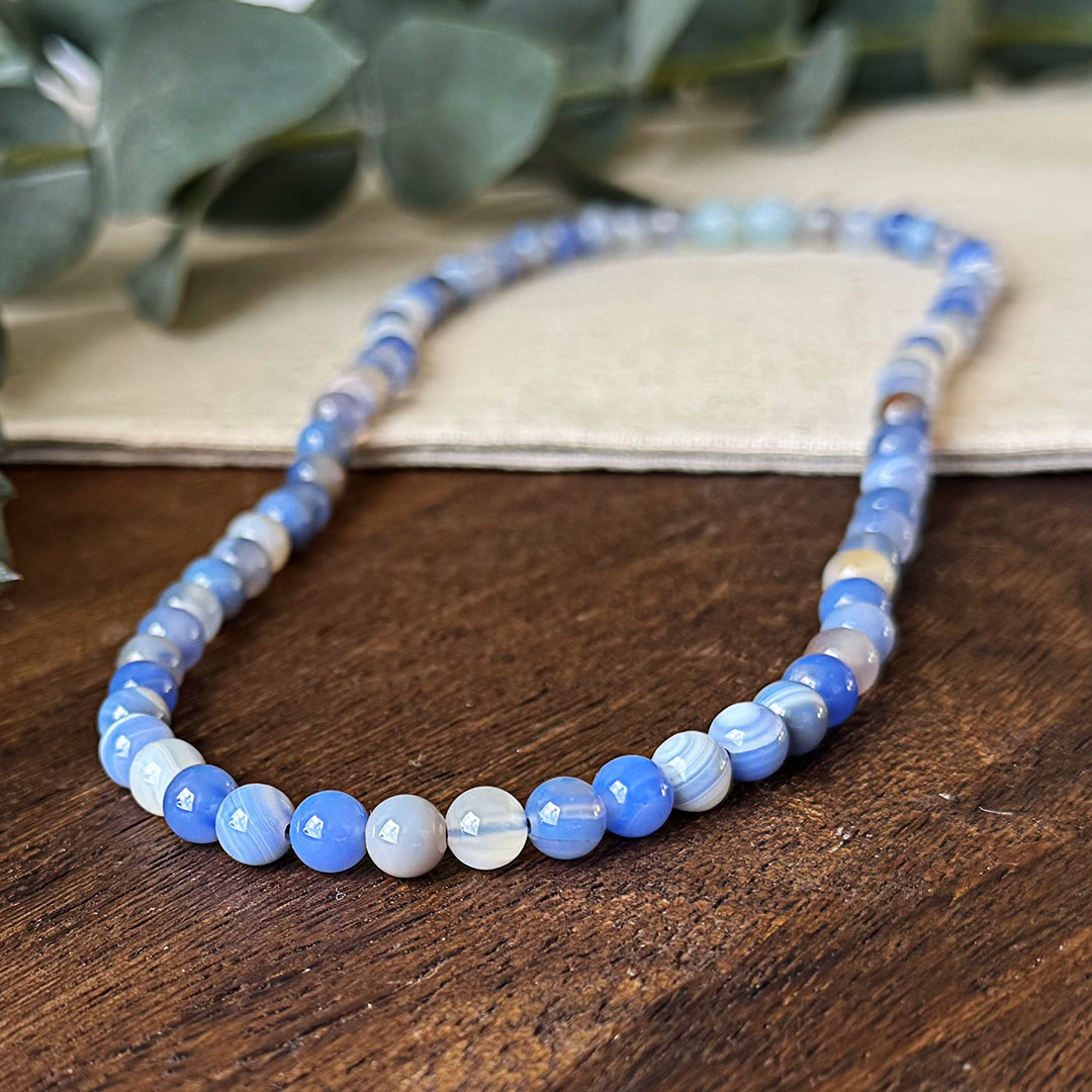 Blue Lace Agate Communication Necklace elegantly displayed on rustic wooden surface. The necklace features uniform round beads in mesmerizing bands of periwinkle blue, soft lavender, and translucent white that capture light beautifully. Each bead shows the stone's natural banding pattern reminiscent of gentle ocean waves. Subtle eucalyptus leaves provide a serene backdrop, complementing the necklace's calming throat chakra energy and highlighting its natural beauty. Perfect for enhancing clear communication
