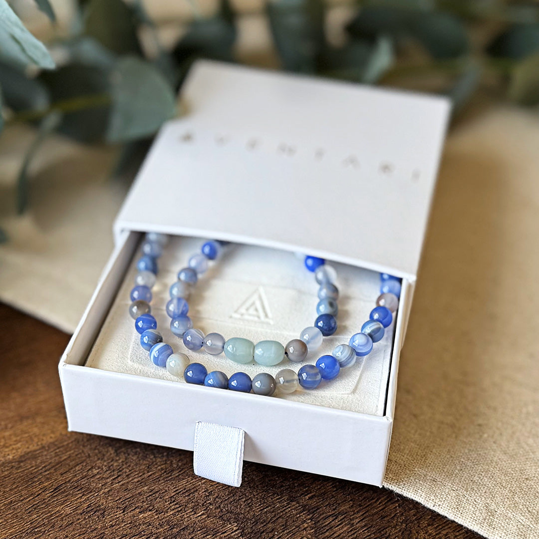 Blue Lace Agate Communication Necklace displayed in an elegant white gift box. The necklace features natural gemstone beads in serene shades of powder blue, lavender, and white, resembling gentle ocean waves. This throat chakra healing crystal jewelry helps promote clear self-expression, confidence, and calm communication while reducing anxiety. Artfully arranged on a cushioned interior with our signature emblem.