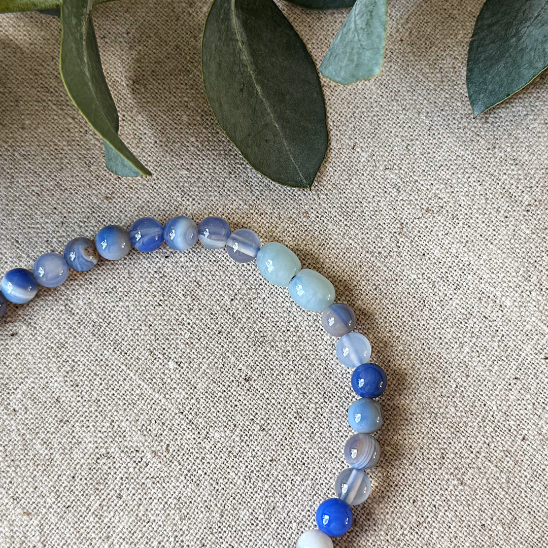 Blue Lace Agate necklace artfully arranged in a gentle curve against natural linen fabric. The polished gemstone beads showcase a mesmerizing gradient of colors from deep violet to pale blue-white, with distinctive banding patterns visible throughout. Fresh eucalyptus leaves frame the top of the image, adding an earthy, organic touch that complements the stone's natural origin. This versatile throat chakra accessory highlights the beautiful translucency and subtle variations in each handcrafted beads.