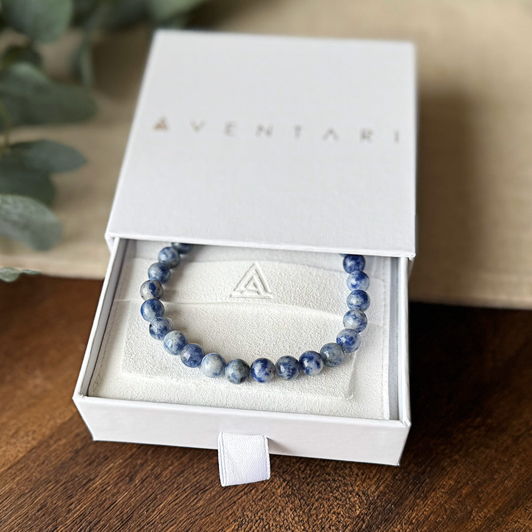 Blue Spot Jasper Serenity Bracelet elegantly presented in a premium Aventari branded gift box. The minimalist white packaging with gold lettering opens to reveal the bracelet nestled on a cream-colored cushion embossed with the brand's triangular logo. The polished gemstone beads showcase their distinctive purple marbling, ready to bring emotional balance and inner strength to their wearer. Perfect for gifting to loved ones needing a touch of tranquility in their daily life.