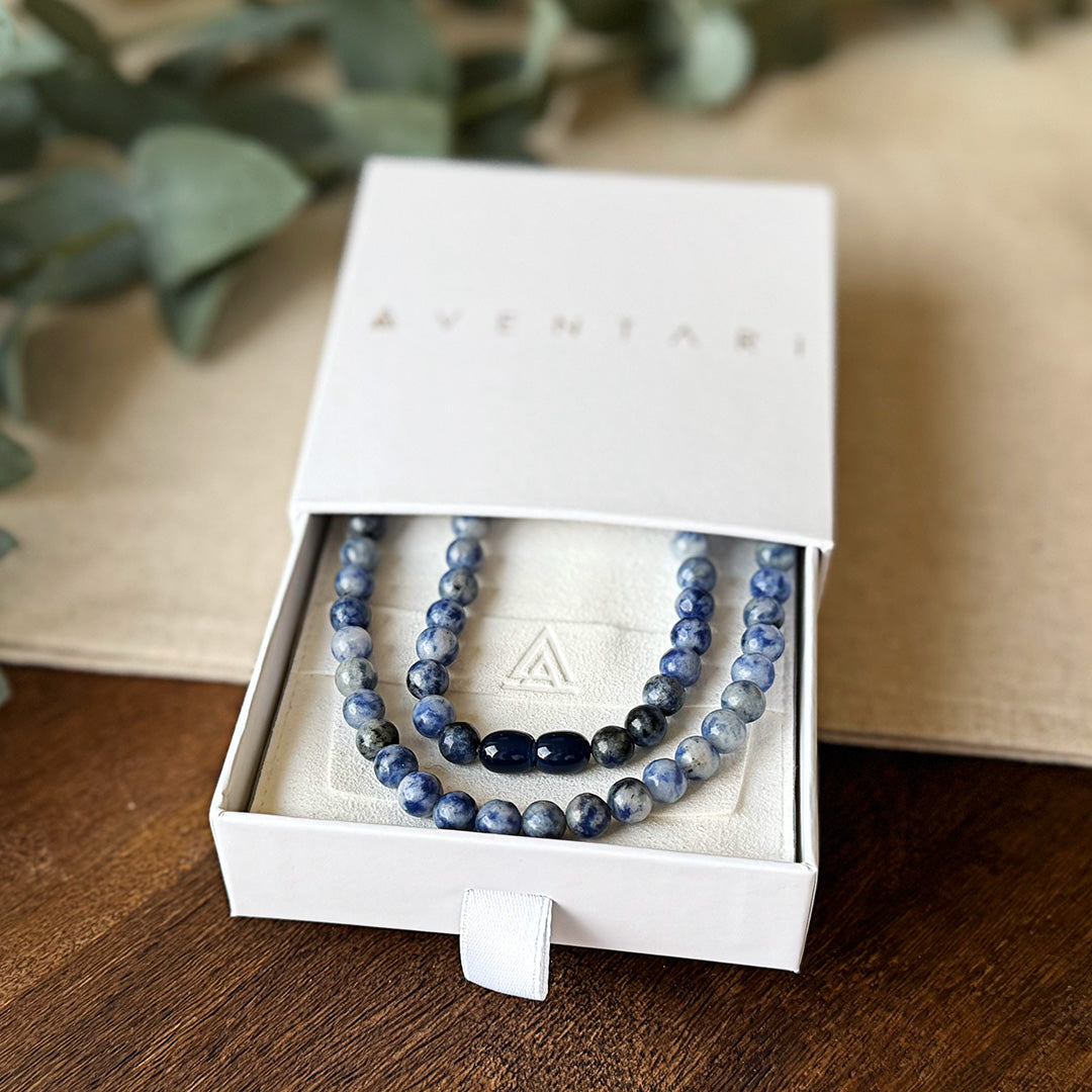 Blue Spot Jasper Serenity Necklace presented in an elegant AVENTARI branded gift box. The premium packaging reveals the necklace nestled on a cream-colored cushion embossed with the brand's triangular logo. Featuring marbled purple and gray beads with accent stones at the center, this piece combines sophistication with natural beauty. Eucalyptus leaves in the background enhance the calming aesthetic, reflecting the necklace's stress-relieving properties.