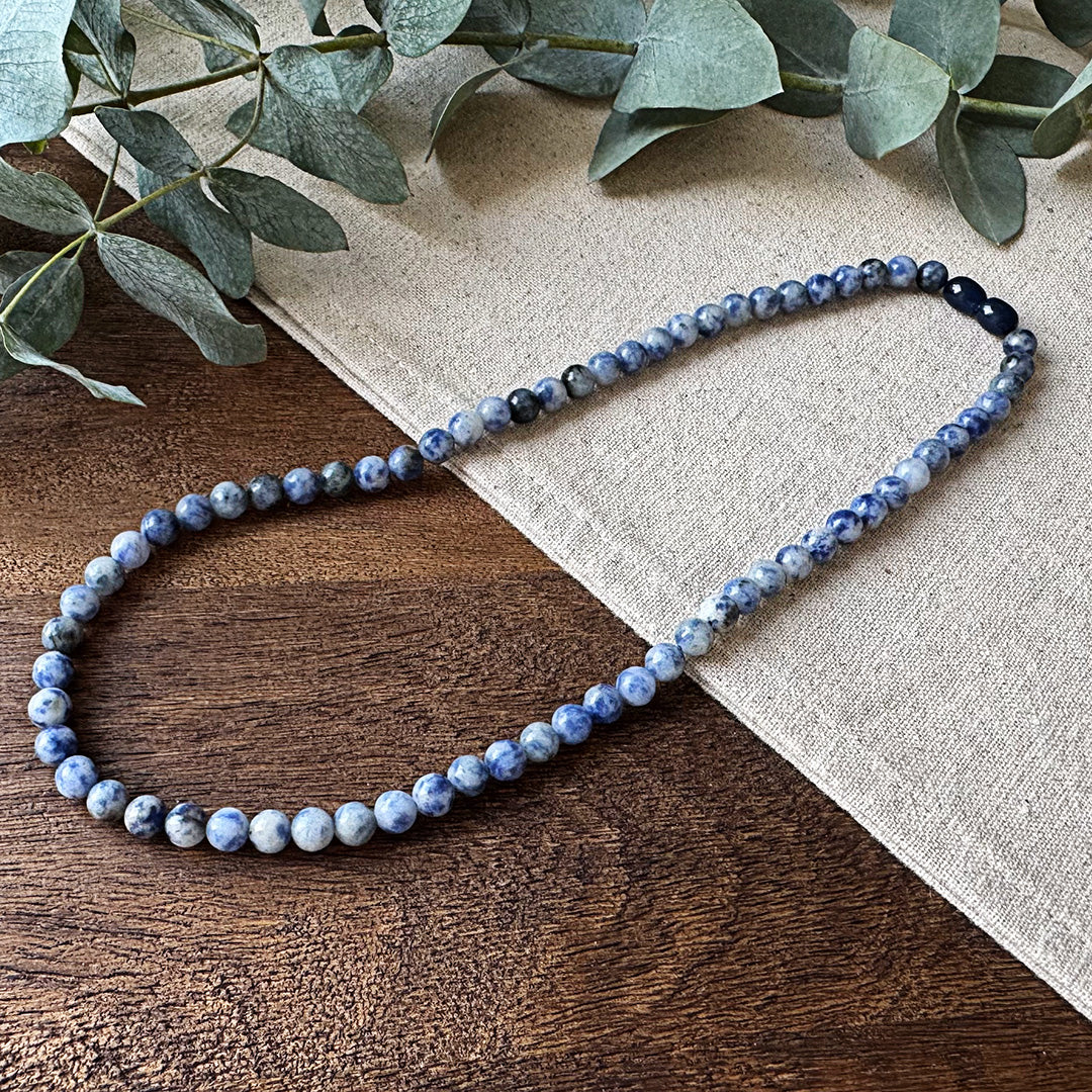 Blue Spot Jasper Serenity Necklace elegantly arranged on rustic wood and natural linen. The polished stone beads showcase distinctive purple and white marbled patterns throughout, creating a striking visual rhythm. Fresh eucalyptus sprigs frame the top corner, adding an earthy, calming element that complements the necklace's tranquil energy properties. Perfect for those seeking balance and inner peace amid life's chaos.
