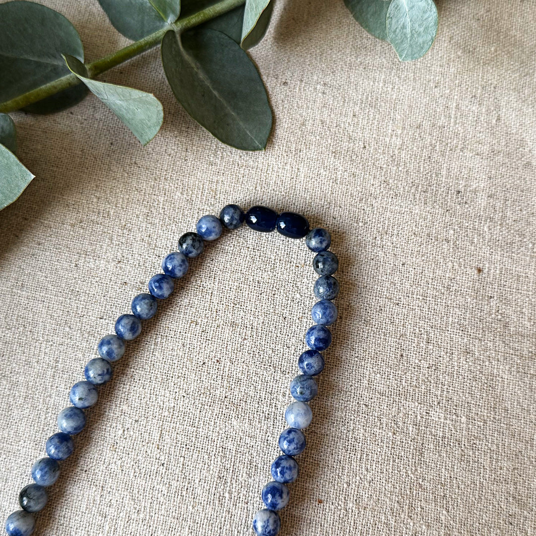 Overhead view of the Blue Spot Jasper Serenity Necklace arranged in a gentle curve on natural linen fabric. The gemstone beads feature stunning purple marbling with distinctive speckled patterns, highlighted by two deeper purple accent stones. Fresh eucalyptus sprigs frame the top portion, complementing the necklace's organic beauty and reinforcing its purpose as a talisman for cultivating inner peace, courage and emotional balance in everyday life.