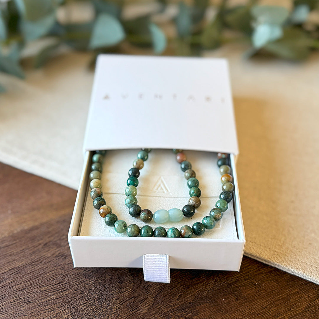 Chrysocolla Wisdom Necklace presented in a premium Aventari branded gift box. The elegant white packaging with gold lettering displays the necklace nestled on a cream-colored cushion embossed with the brand's triangular logo. The polished gemstone beads exhibit their beautiful spectrum of greens and blues with distinctive pale blue amazonite accent beads. Each stone's unique patterning reflects the necklace's connection to nature and its calming, intuition-enhancing properties.