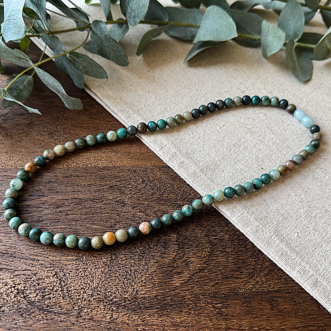 Chrysocolla Wisdom Necklace elegantly arranged on rustic wood and natural linen fabric. The polished gemstone beads showcase a stunning array of earthy greens, teals, and blues with natural variations and marbling throughout. Two pale blue amazonite accent beads create a focal point in the design. Fresh eucalyptus leaves frame the top corner, complementing the necklace's nature-inspired palette. This versatile piece combines earthy elegance with the stone's reputed properties of tranquility, wisdom, and emo