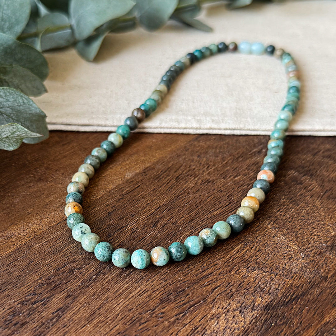 Close-up of the Chrysocolla Wisdom Necklace artfully displayed on natural wood and linen. The carefully selected beads showcase nature's artistry with their variegated tones of forest green, teal, and earth tones, punctuated by luminous pale blue amazonite accent beads. Soft-focus eucalyptus leaves create a serene backdrop, emphasizing the harmony between the stone's natural beauty and its traditional associations with tranquility, communication, and emotional balance.