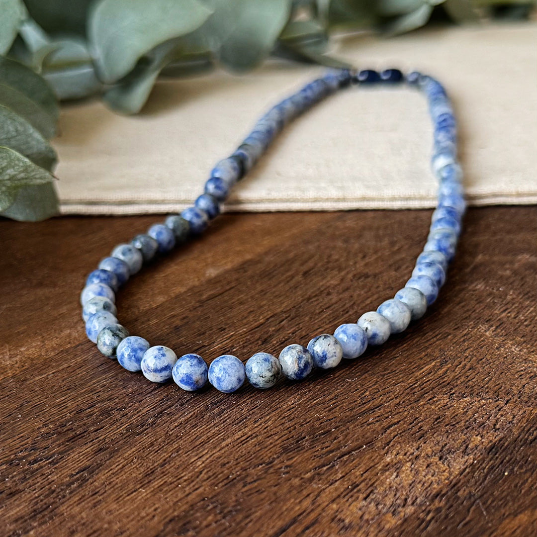 Close-up of the Blue Spot Jasper Serenity Necklace gracefully draped on rich wooden surfaces and natural linen. The polished beads display captivating variations of purple marbling against white and gray backgrounds, showcasing nature's artistry in each uniquely patterned stone. Soft-focus eucalyptus leaves create a serene backdrop, emphasizing the necklace's calming influence and connection to nature's tranquility.
