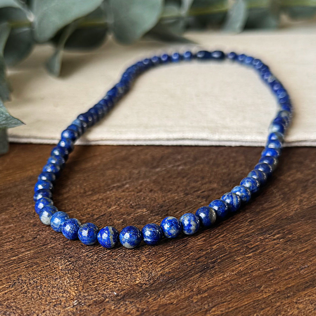 Close-up of the Lapis Lazuli Protection Necklace artfully displayed on natural wood and linen. The uniform polished beads showcase their deep, intense blue color with natural gold pyrite inclusions that catch the light beautifully. Soft-focus eucalyptus leaves create a serene backdrop, highlighting the necklace's connection to nature while emphasizing its properties as a stress-relieving and truth-enhancing stone. The vivid blue color stands out dramatically against the neutral background.