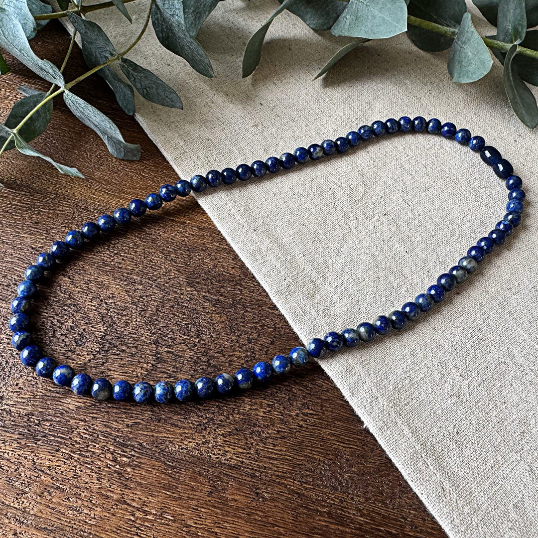 Lapis Lazuli Protection Necklace elegantly arranged on rustic wood and natural linen fabric. The polished gemstone beads showcase a stunning deep royal blue color with characteristic gold pyrite flecks that shimmer in the light. Fresh eucalyptus sprigs frame the upper corner, complementing the necklace's natural beauty while emphasizing its protective and calming qualities. Each bead displays the stone's distinctive rich coloration, creating a striking piece that combines visual appeal with traditional prop
