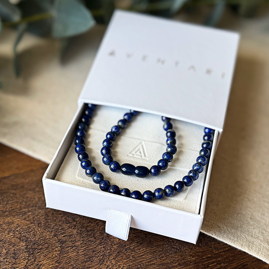 Lapis Lazuli Protection Necklace presented in a premium Aventari branded gift box. The elegant white packaging with gold lettering displays the necklace nestled on a cream-colored cushion embossed with the brand's triangular logo. The vibrant cobalt blue beads feature natural gold-flecked inclusions, with two oval accent beads creating a focal point in the design. The necklace's rich color symbolizes wisdom, truth and protection against negative energies, making it both a luxurious gift and powerful talisma