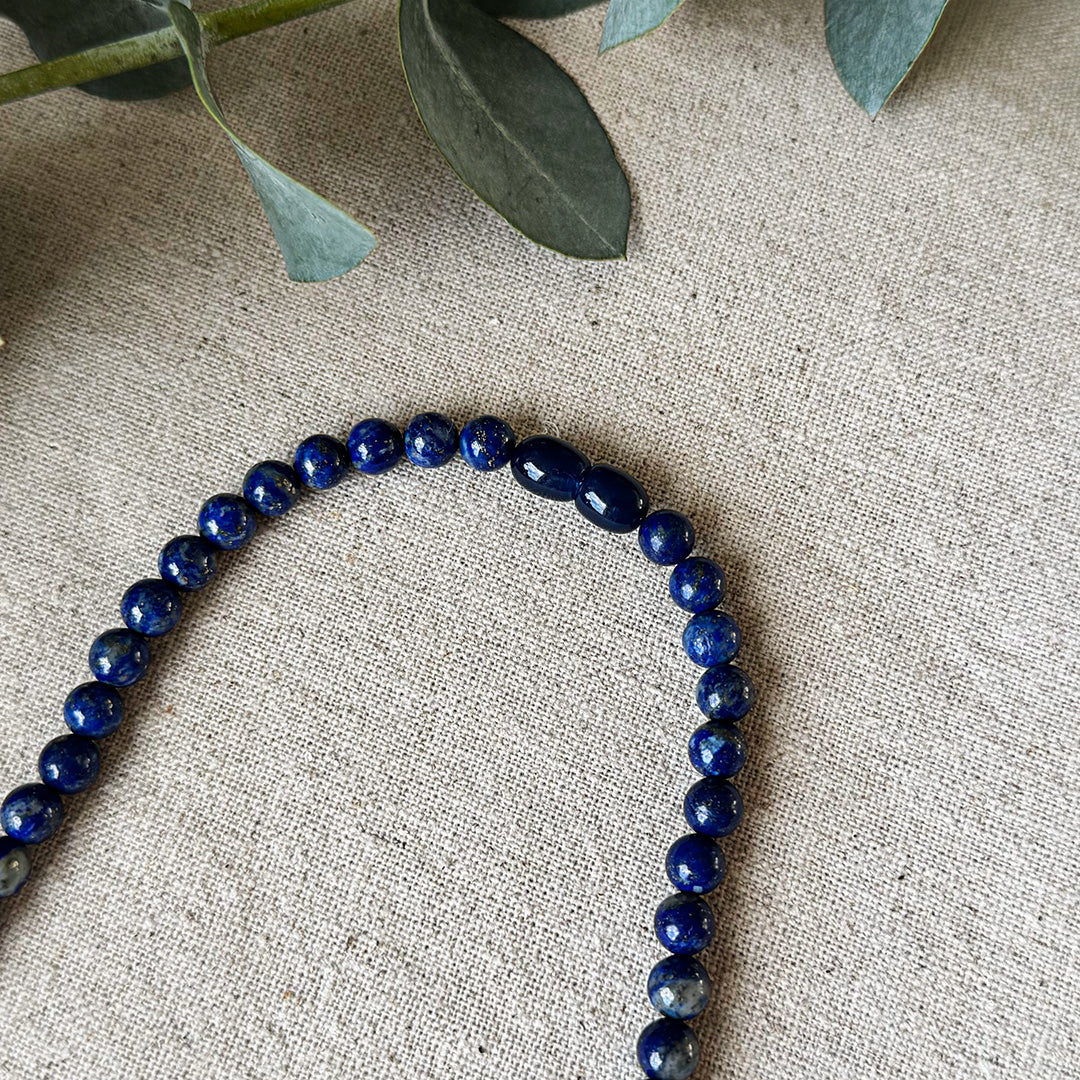 Detail view of the Lapis Lazuli Protection Necklace on natural linen fabric with eucalyptus accents. The image highlights the gemstone's signature deep blue color with golden pyrite flecks and two oval-shaped accent beads that create visual interest. Each polished stone reveals subtle variations in the distribution of gold inclusions against the rich cobalt background. This powerful piece serves as both a striking accessory and a wearable talisman for enhancing confidence, truth-speaking, and protection.