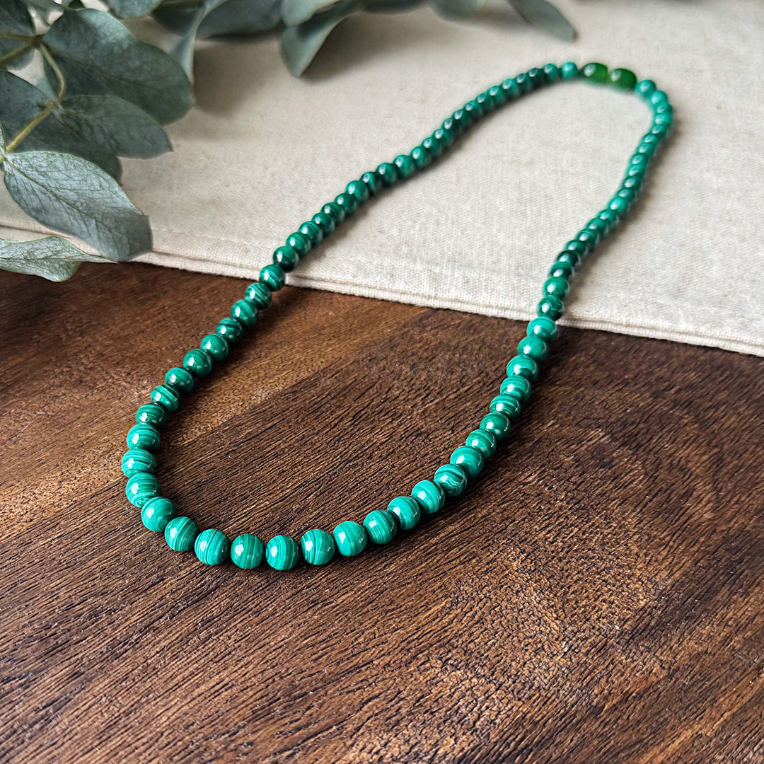 Close-up of the Malachite Transformation Necklace artfully displayed on natural wood and linen. The polished beads reveal their hypnotic green striations that vary from light to dark, creating a mesmerizing visual rhythm throughout the piece. Soft-focus eucalyptus leaves create a serene backdrop, complementing the necklace's earth tones while highlighting its properties as a stone of transformation, emotional balance, and personal growth.