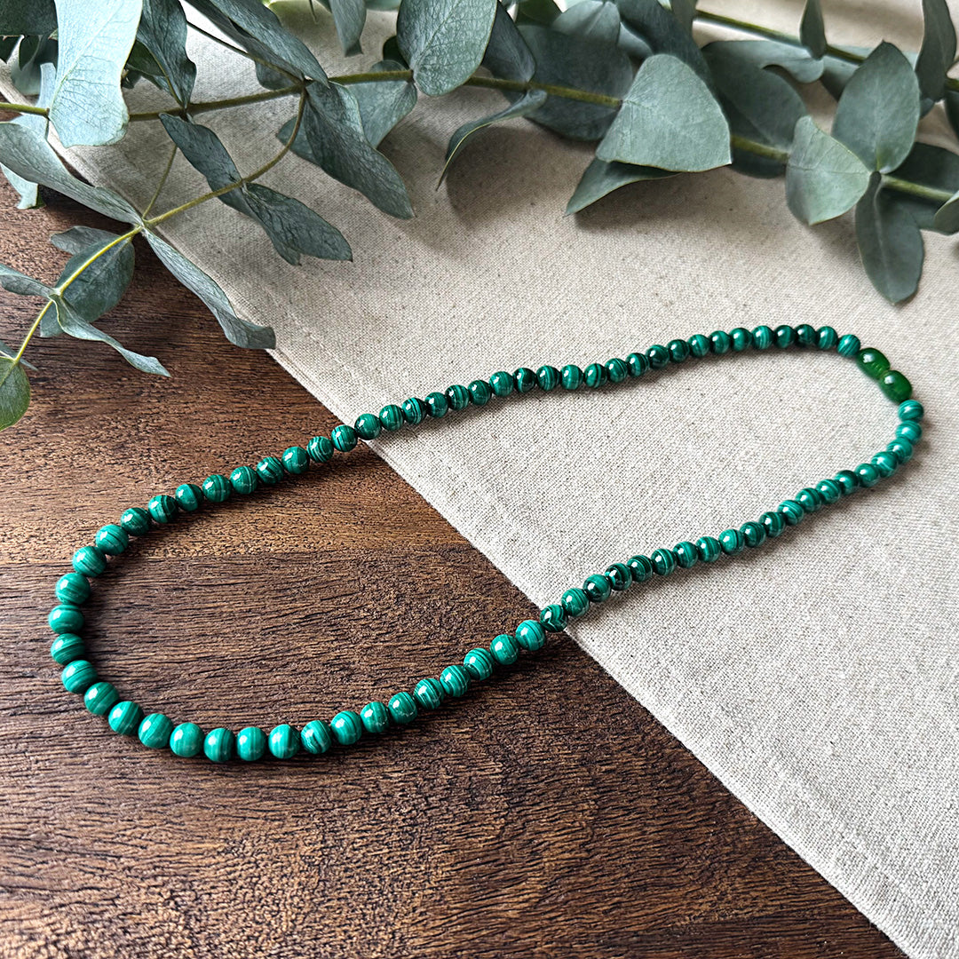 Malachite Transformation Necklace elegantly arranged on rustic wood and natural linen fabric. The polished gemstone beads showcase mesmerizing green banding patterns with distinctive striations that swirl throughout each stone. The necklace rests partially on wood and partially on linen, creating visual interest that highlights the gemstone's natural beauty. Fresh eucalyptus leaves frame the upper portion, complementing the necklace's verdant tones.