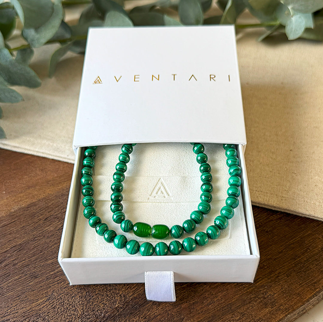 Malachite Transformation Necklace presented in a premium Aventari branded gift box with gold lettering. The elegant white packaging displays the necklace nestled on a cream-colored cushion embossed with the brand's triangular logo. The vibrant green beads exhibit their characteristic banding patterns, with two oval accent beads creating a focal point. Each stone's unique swirling patterns resemble natural fingerprints, showcasing the gemstone's distinctive beauty.