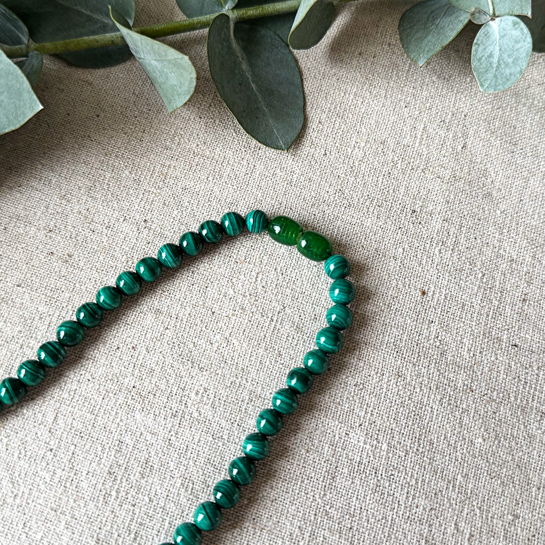 Detail view of the Malachite Transformation Necklace on natural linen fabric with eucalyptus accents. The image highlights the distinctive green oval accent beads and the stone's signature swirling patterns that resemble natural topography. Each polished bead displays unique striations ranging from forest to mint green, capturing light beautifully across their smooth surfaces. This transformative piece serves as both a striking accessory and a wearable talisman for encouraging positive change.