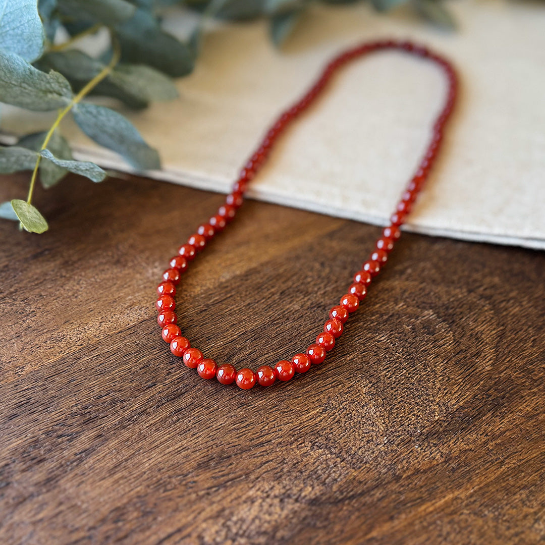 Close-up of the Red Agate Courage Necklace artfully displayed against rustic wood and natural linen with soft-focus eucalyptus leaves. The translucent crimson beads showcase their natural variation and depth, with light dancing across their polished surfaces. Each bead carries the stone's energetic properties of protection, strength, and passionate determination—a wearable talisman designed to enhance confidence and courage throughout daily challenges.