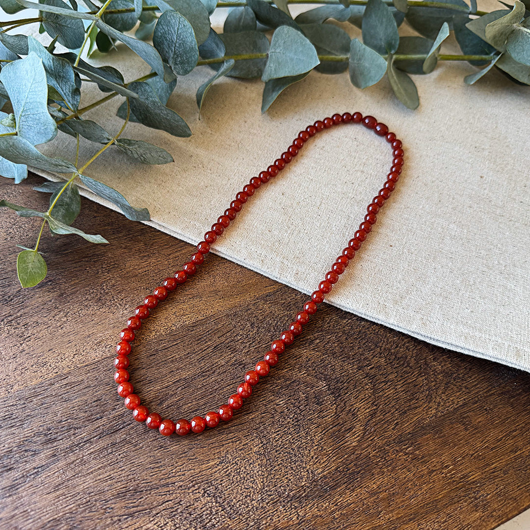 Red Agate Courage Necklace elegantly arranged on a natural wood surface and linen fabric. The polished gemstone beads showcase a rich crimson color with exceptional clarity and shine, forming a complete circle that symbolizes continuous strength and protection. Fresh eucalyptus sprigs frame the top of the image, creating an earthy backdrop that complements the stone's grounding energy. Perfect for those seeking to amplify confidence, creativity, and inner fire in their daily life.