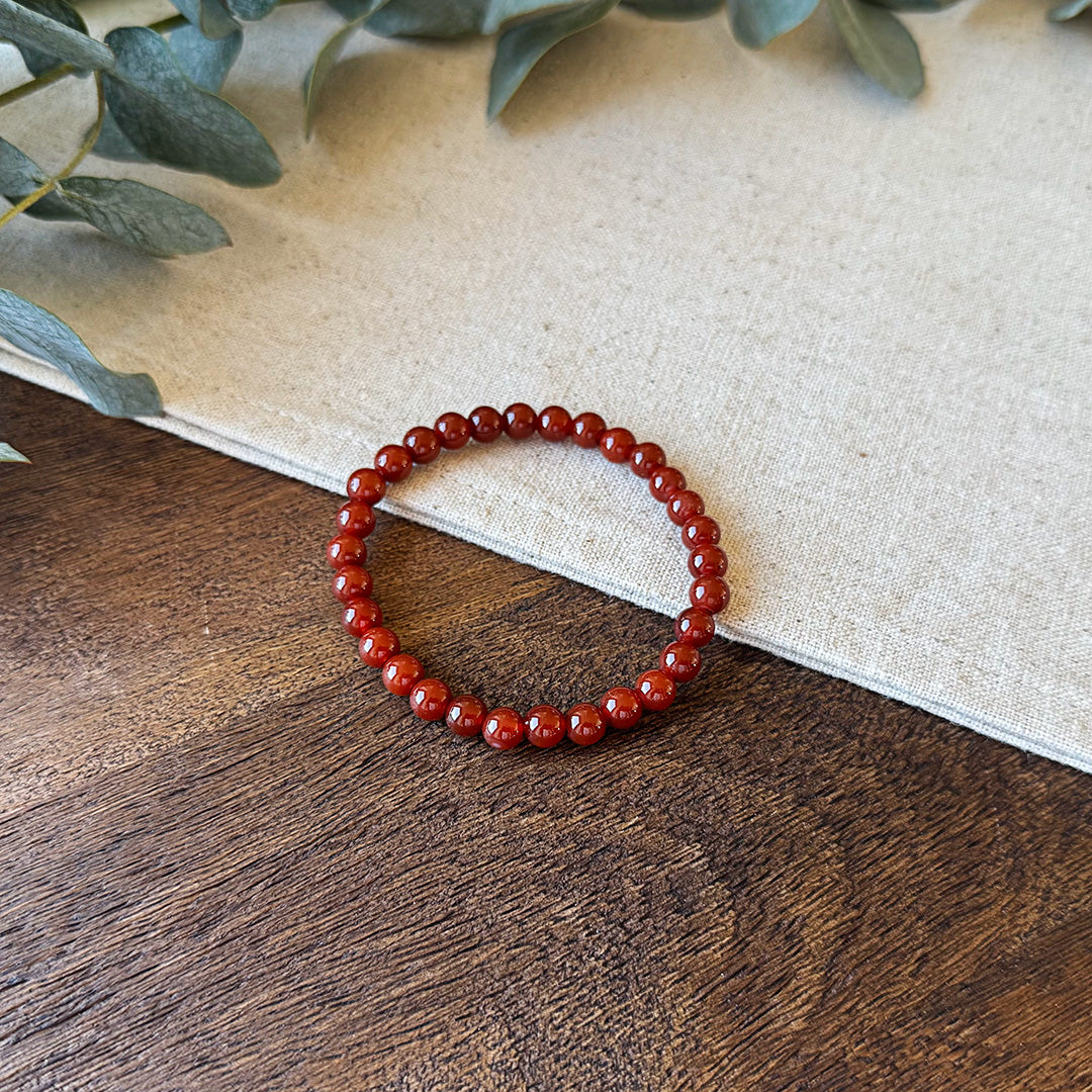 Red Agate Courage Bracelet formed in a perfect circle on rustic wood and natural linen fabric. The bracelet features uniform polished beads in a rich, deep crimson color that catches the light with a glossy sheen. Each bead's translucent quality reveals subtle depth variations, symbolizing inner strength and passionate energy. Soft eucalyptus leaves frame the upper corner, adding an earthy element that complements the stone's grounding properties. This wearable talisman is designed to amplify confidence and