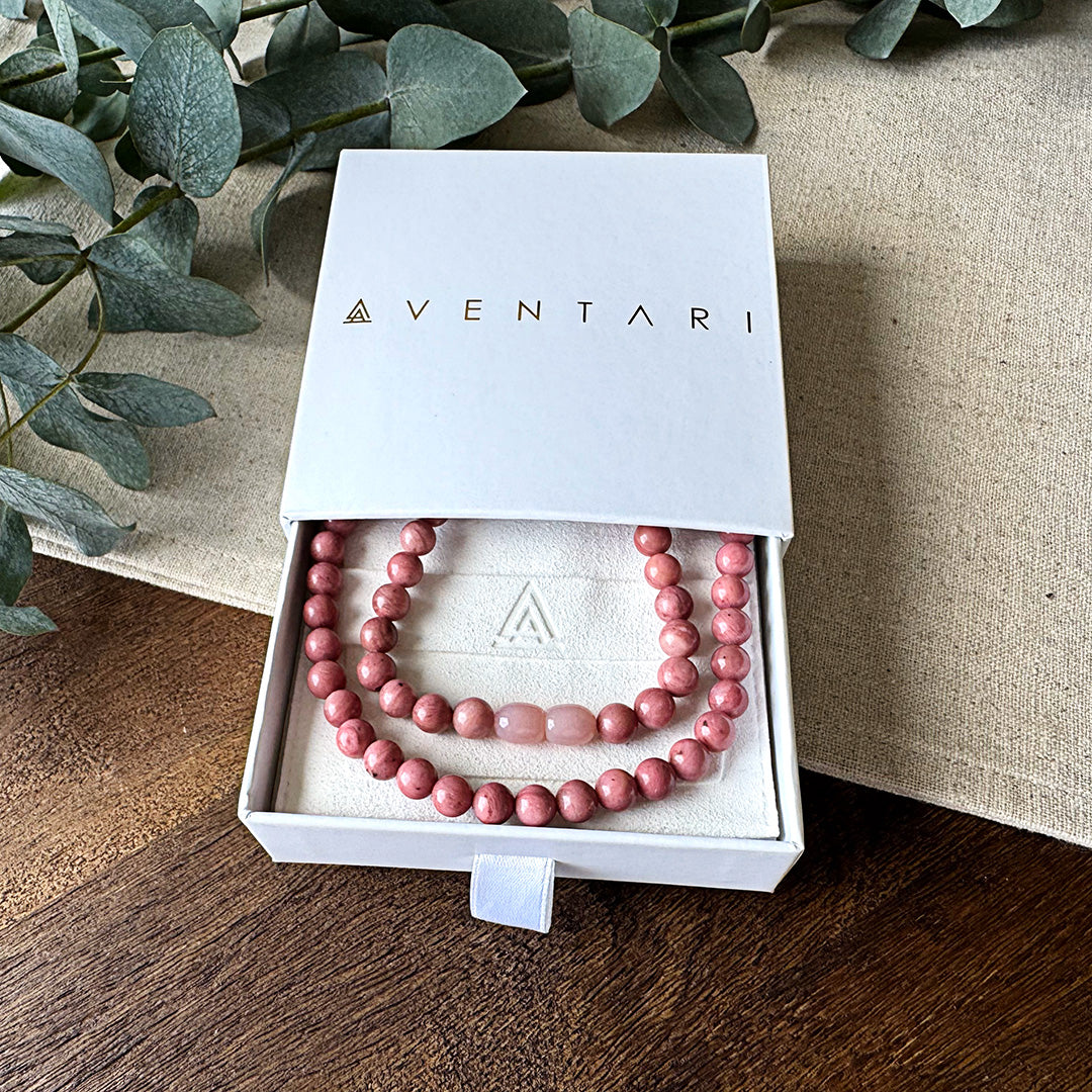 Rhodonite Healing Necklace presented in a premium Aventari branded gift box. The elegant white packaging with gold lettering displays the necklace nestled on a cream-colored cushion embossed with the brand's triangular logo. The beautiful pink-rose colored beads exhibit their natural variations with two lighter oval accent beads creating a focal point. Each gemstone showcases the stone's characteristic matrix patterns, symbolizing emotional healing and heart chakra balance. Perfect for gifting.