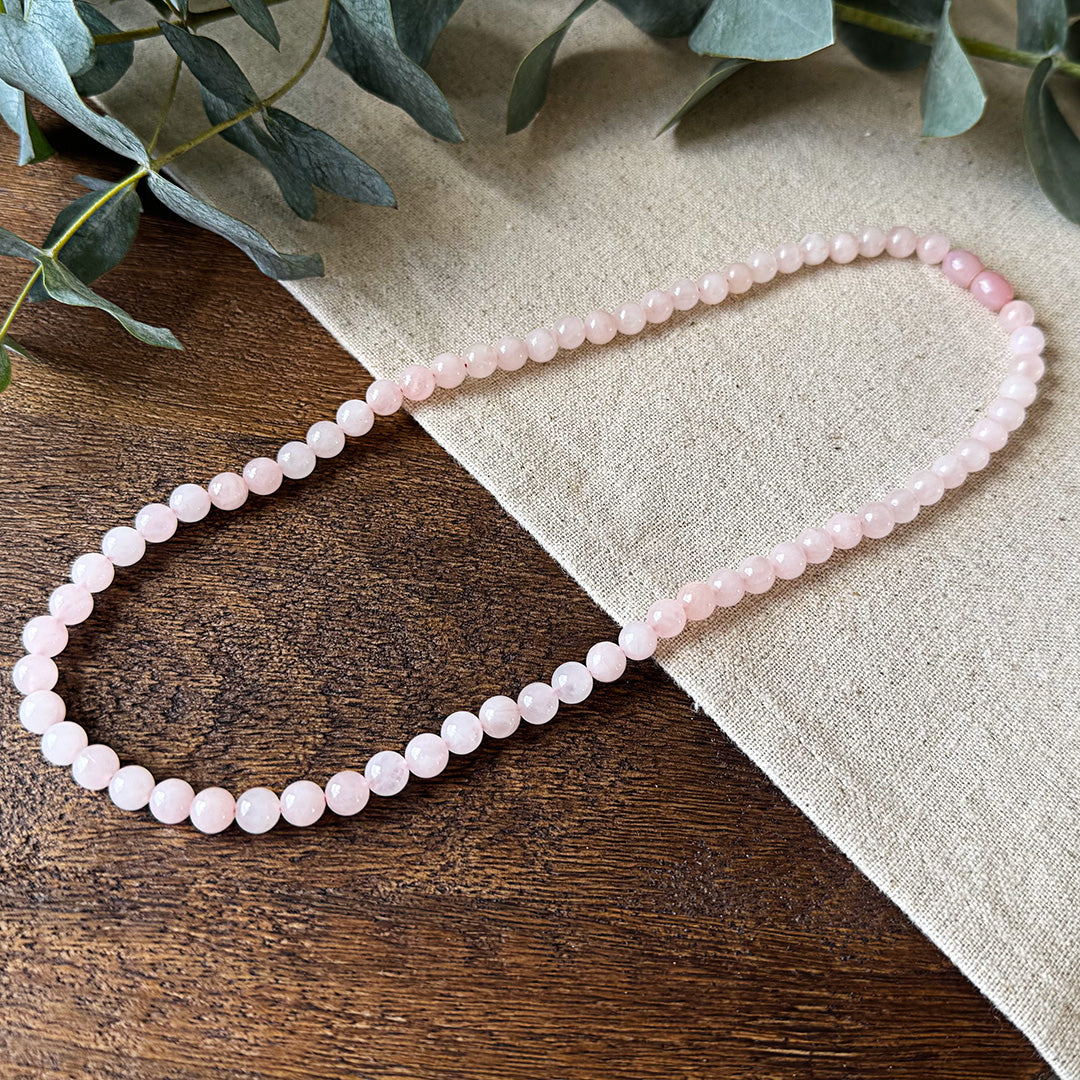 Rose Quartz Love Necklace elegantly arranged on rustic wood and natural linen fabric. The polished gemstone beads showcase a delicate pink color with subtle translucency that catches the light beautifully. The necklace features soft rose-colored round beads with two slightly darker oval accent beads at the center. Fresh eucalyptus leaves frame the upper corner, creating an earthy contrast to the necklace's gentle feminine energy. This heart chakra stone is known for its properties of unconditional love.