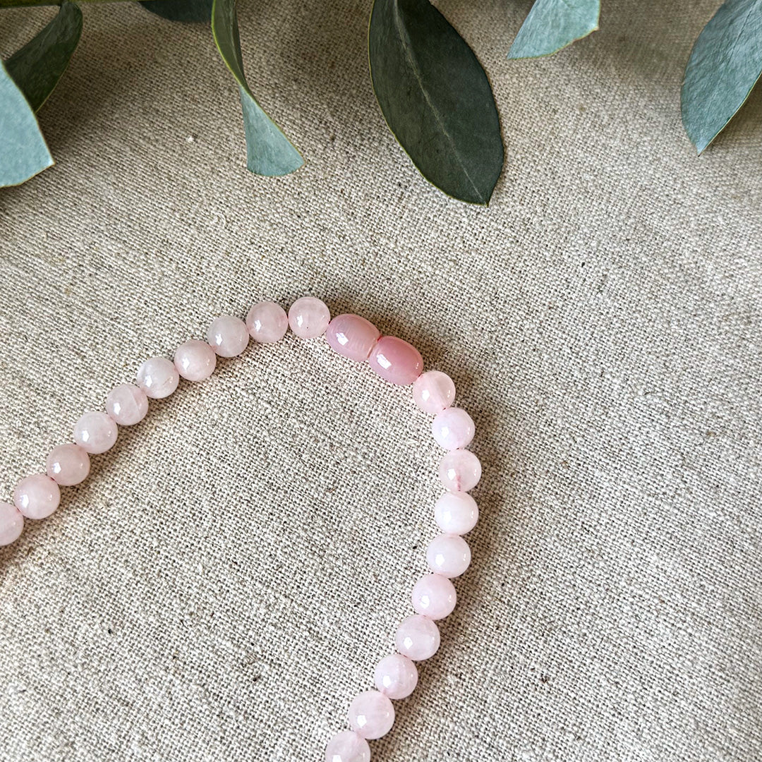 Detail view of the Rose Quartz Love Necklace on natural linen fabric with eucalyptus accents. The image highlights the distinctive oval clasp and the stone's signature soft pink color that varies slightly throughout each polished bead. Each stone reflects light with a gentle luminosity, emphasizing the necklace's connection to heart-centered energy. This beautiful piece serves as both a stylish accessory and a wearable talisman for attracting love, fostering self-acceptance, and promoting healing.