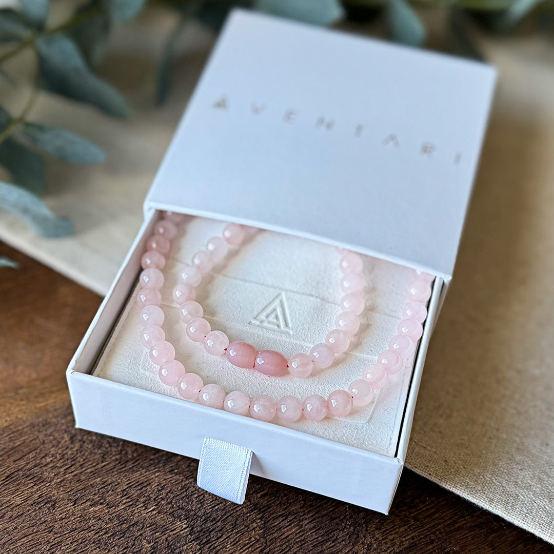 Rose Quartz Love Necklace presented in a premium Aventari branded gift box. The elegant white packaging with gold lettering displays the necklace nestled on a cream-colored cushion embossed with the brand's triangular logo. The soft pink gemstone beads exhibit a luminous quality with two oval accent beads creating a focal point. Each bead showcases the stone's characteristic gentle pink hue, symbolizing universal love and heart healing. Perfect for gifting to a loved one.