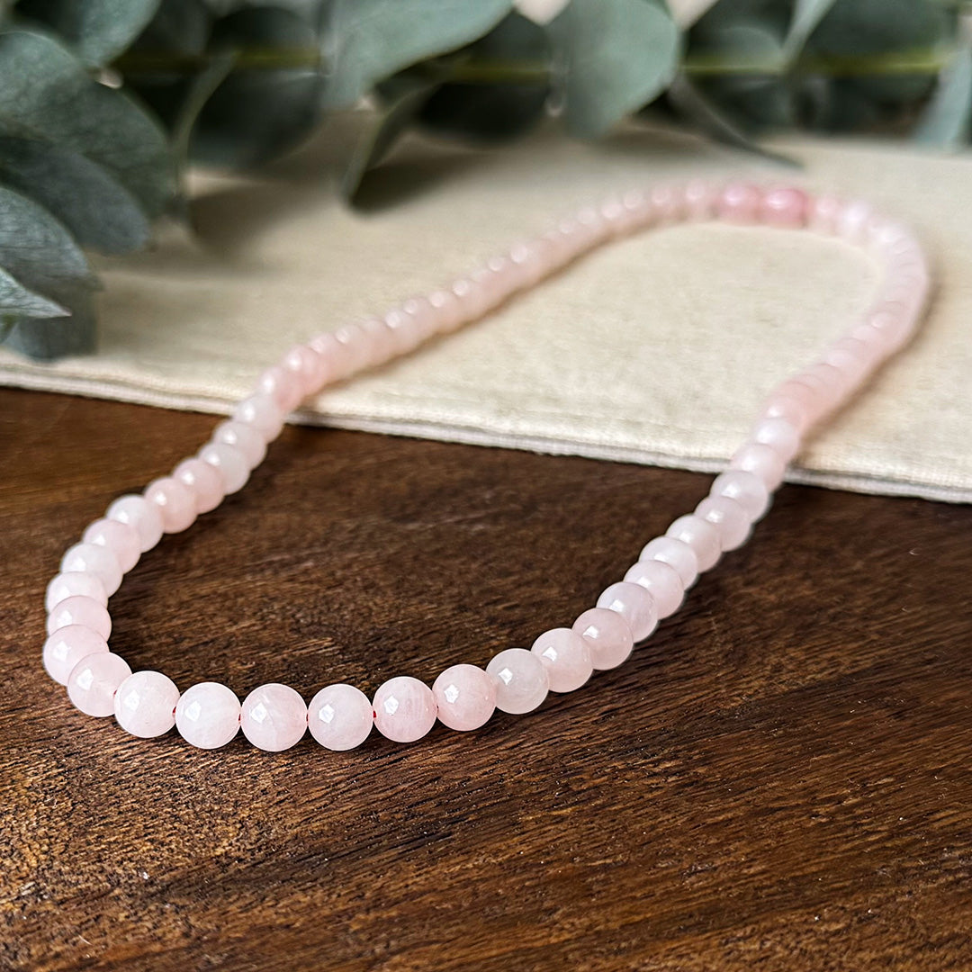 Close-up of the Rose Quartz Love Necklace artfully displayed on natural wood and linen. The uniform polished beads reveal their gentle pink coloration with subtle internal variations that create a soft, romantic glow. Soft-focus eucalyptus leaves create a serene backdrop, complementing the necklace's nurturing energy while highlighting its properties as a stone of unconditional love, emotional healing, and heart chakra alignment.
