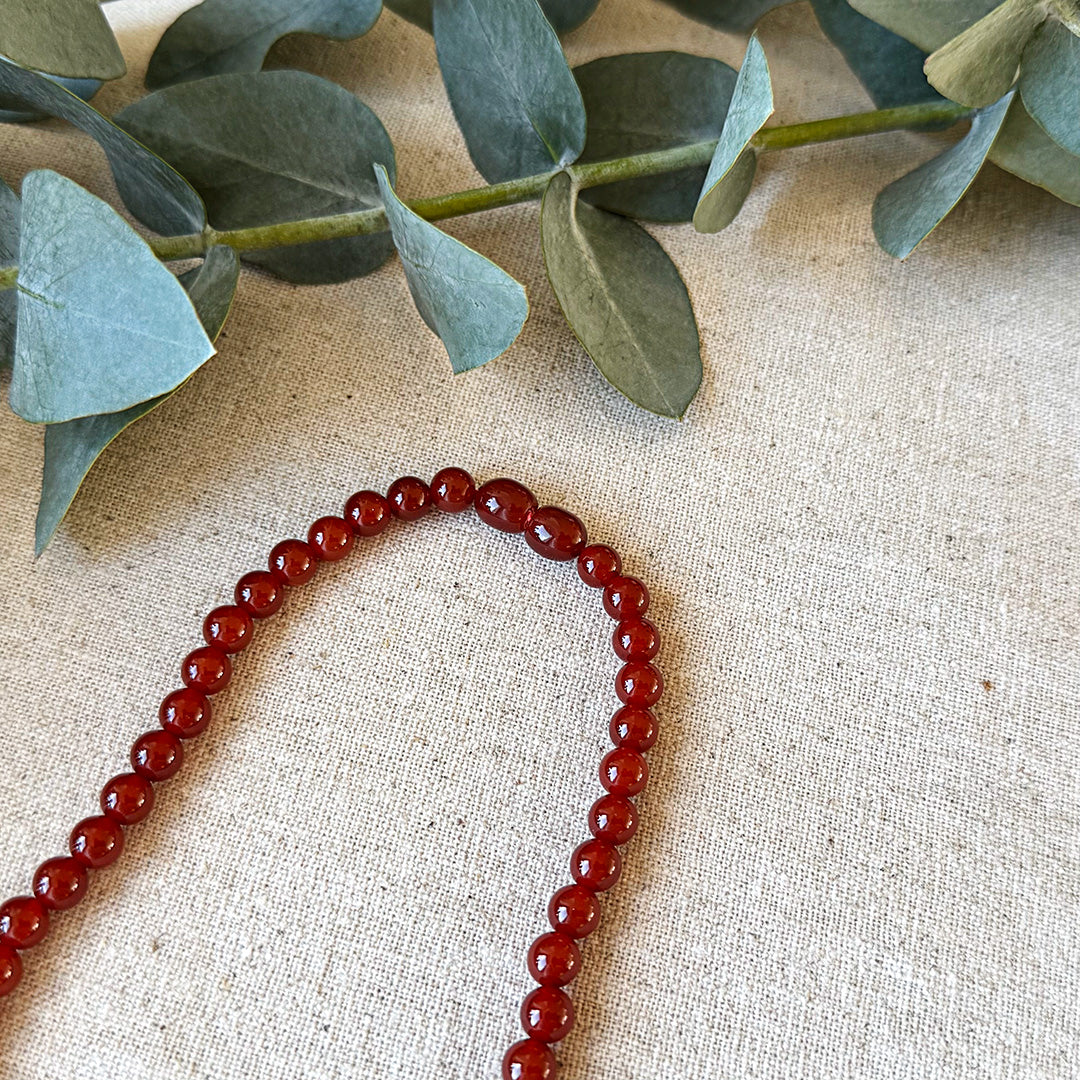 Overhead view of the Red Agate Courage Necklace arranged in a gentle curve on natural linen fabric. The rich, deep red beads display their remarkable consistency in color and polish, creating a powerful visual statement against the neutral background. Fresh eucalyptus leaves frame the top portion, symbolizing growth alongside the necklace's energy for courage. This fiery gemstone accessory serves as both a striking fashion piece and a daily reminder of one's inner strength and bold potential.