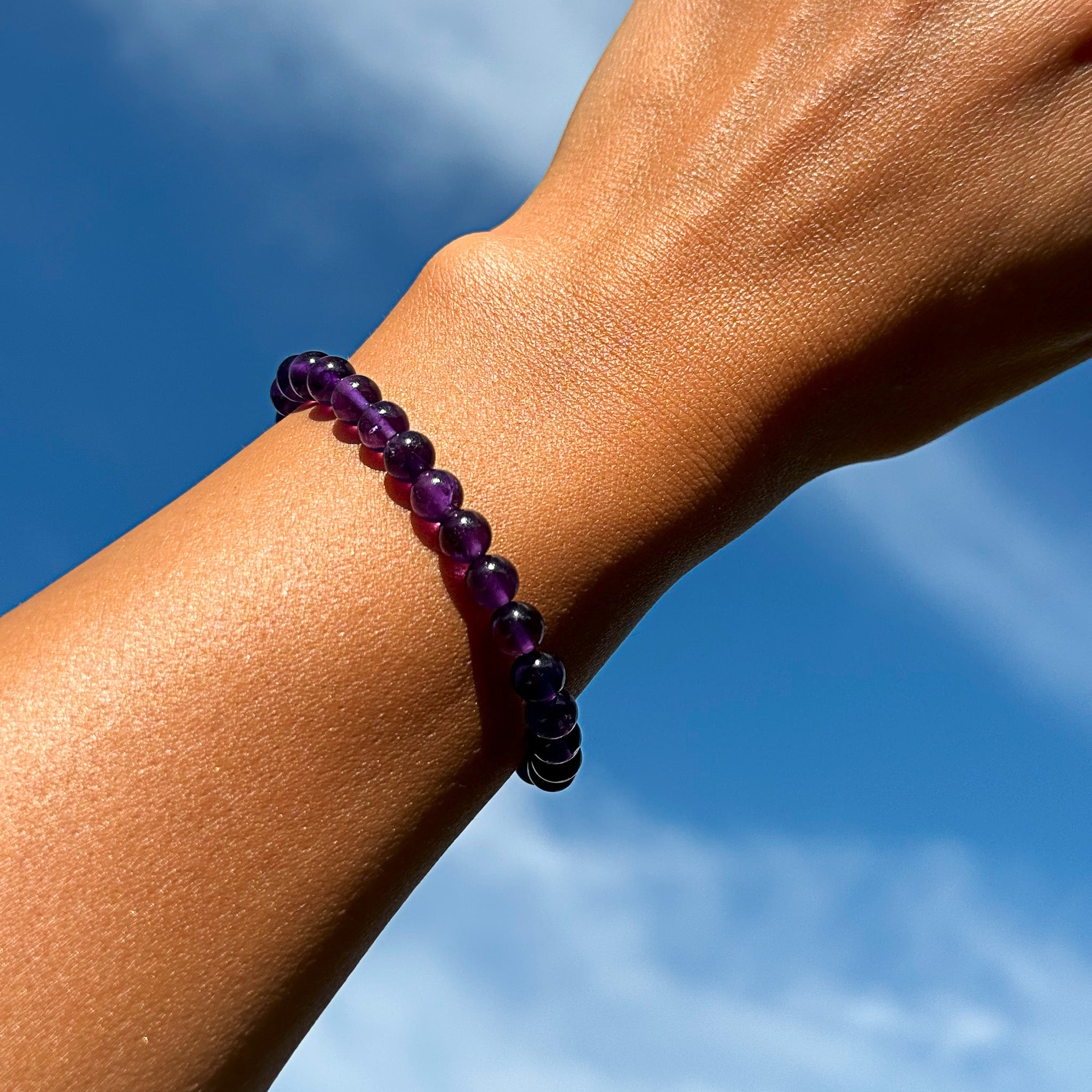 Close-up of the Aventari Amethyst Bracelet featuring vibrant purple beads worn on a wrist against a bright blue sky background.