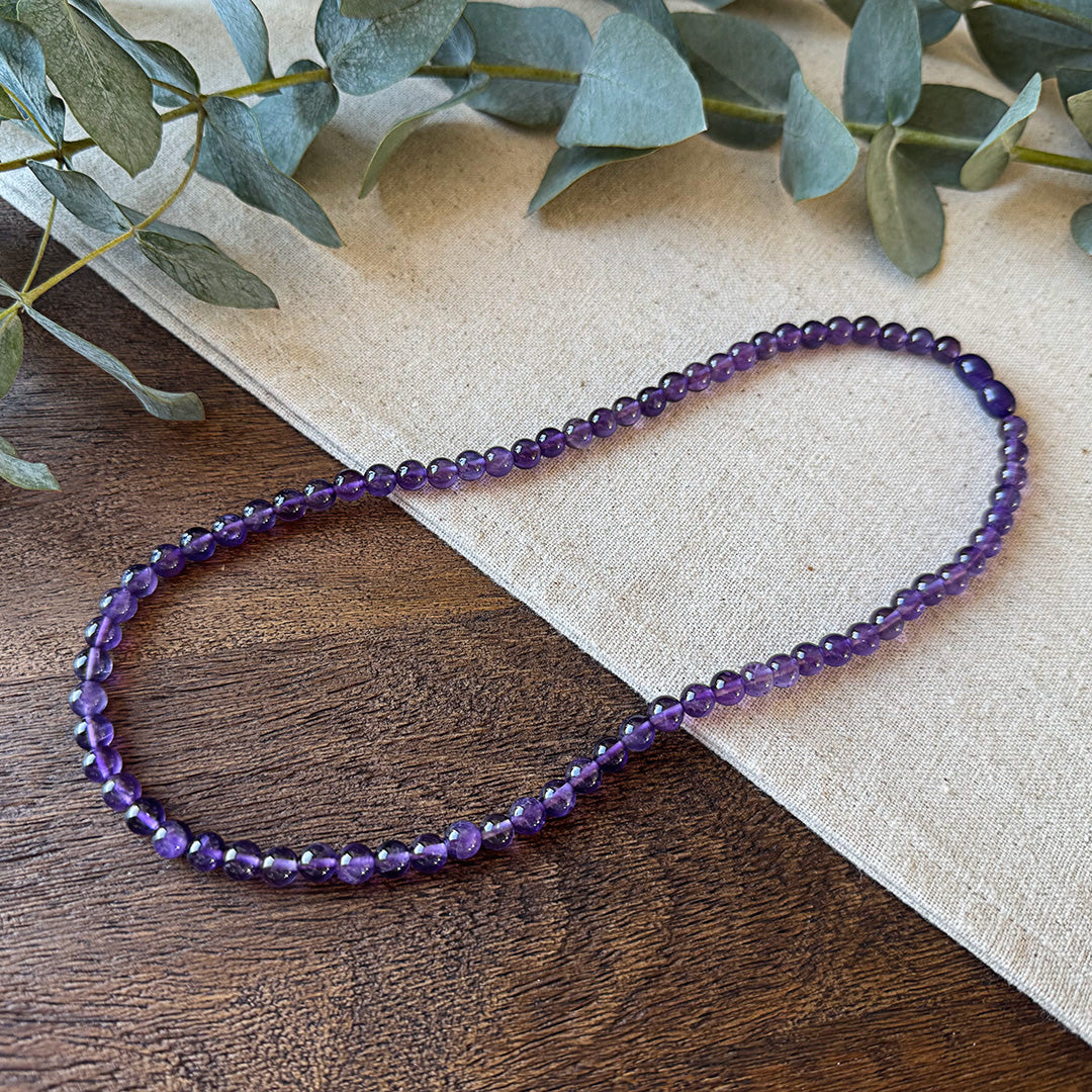 Aventari Amethyst Spiritual Power Necklace with deep purple beads laid flat on a natural wood and linen surface with eucalyptus.