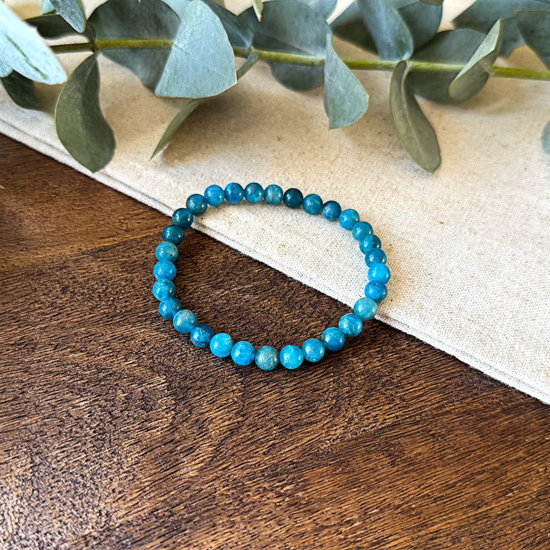 Aventari Apatite Intellect Bracelet featuring blue-green beads laid flat on a natural wood and linen surface with eucalyptus accents.