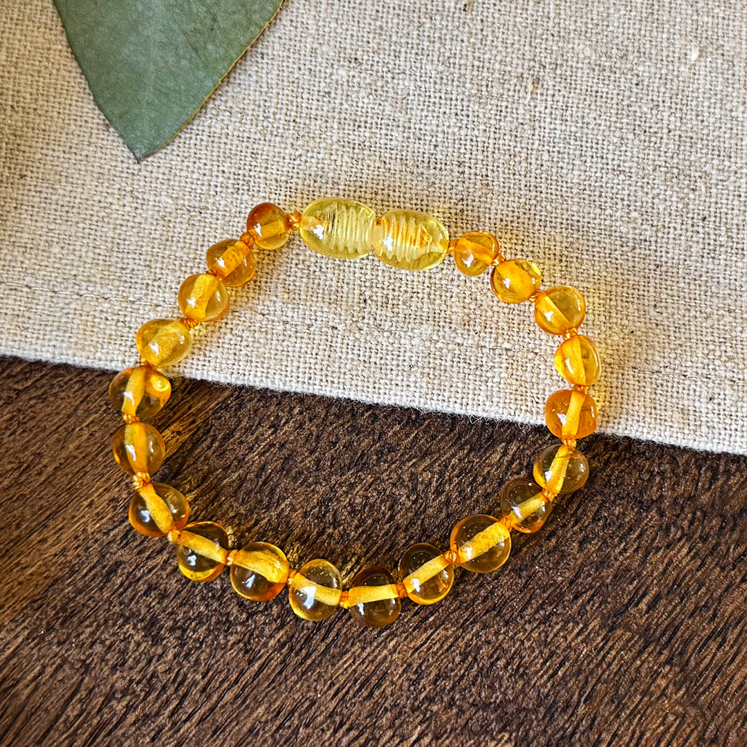Aventari handcrafted Baltic Amber Soothing Bracelet with golden beads and screw clasp resting on natural wood and linen with eucalyptus leaves.