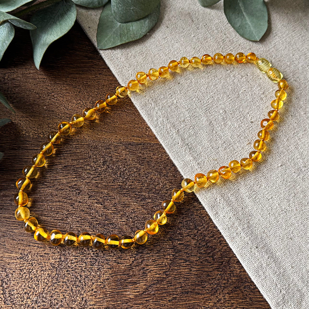 Aventari Baltic Amber Soothing Necklace featuring warm, golden beads laid flat in a circle on a natural wood and linen background with eucalyptus accents.