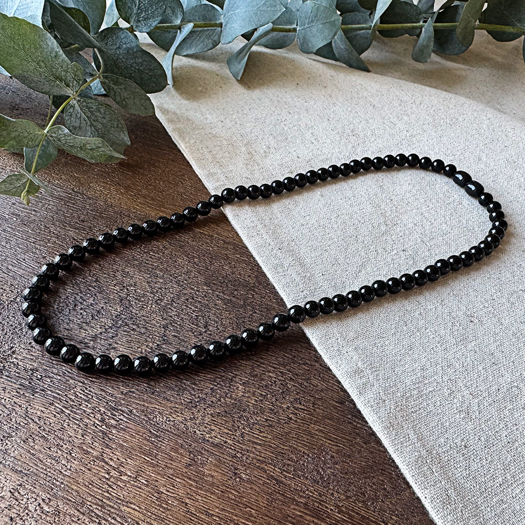 Aventari Black Onyx Focus Necklace featuring polished black beads laid flat on a natural wood and linen surface with eucalyptus.