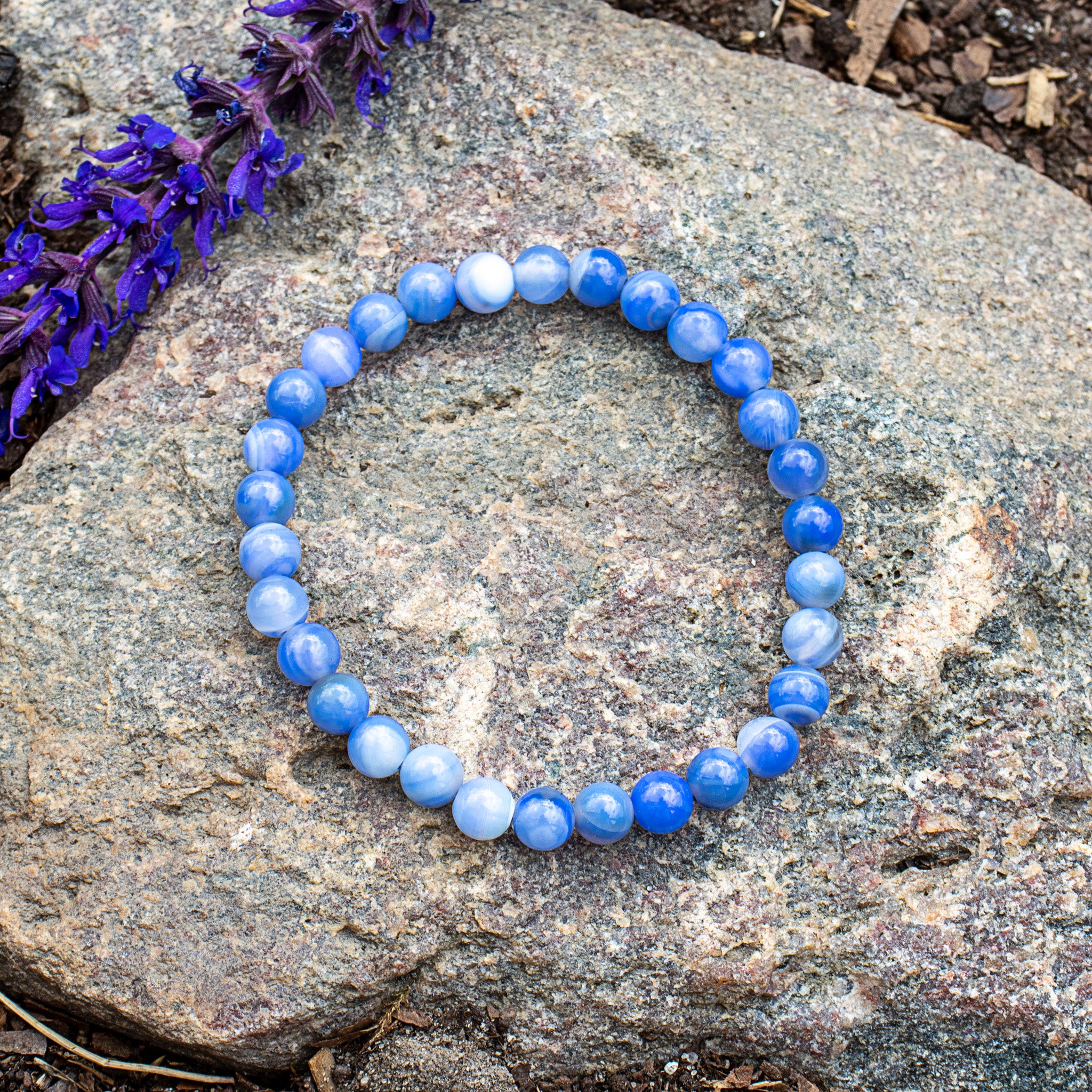 Aventari Blue Lace Agate Bracelet with soothing blue bands displayed on a granite rock next to purple flowers outdoors.