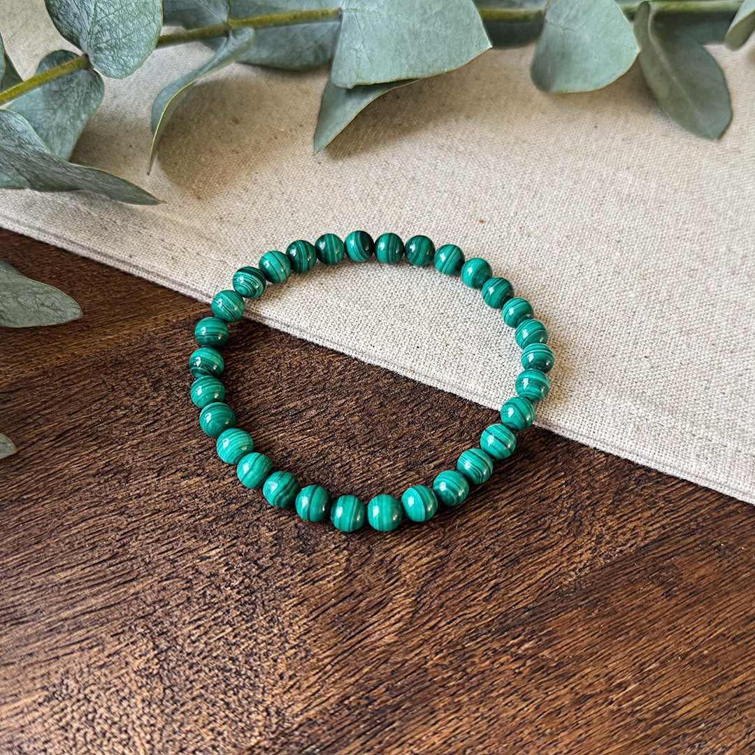 Aventari Malachite Transformation Bracelet featuring green banded beads resting on a natural wood and linen surface.