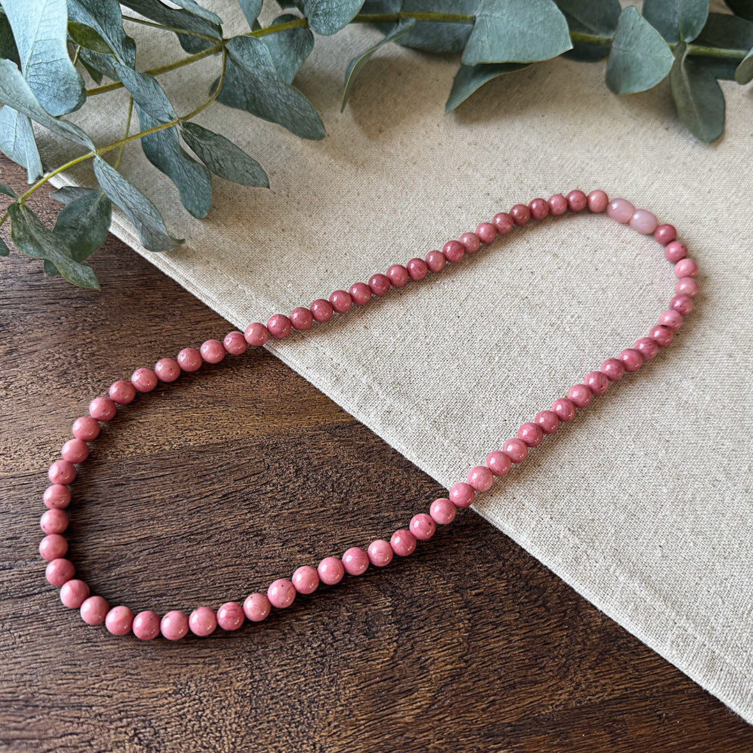 Aventari Rhodonite Healing Necklace featuring dusty pink beads laid flat on a natural wood and linen surface with eucalyptus.