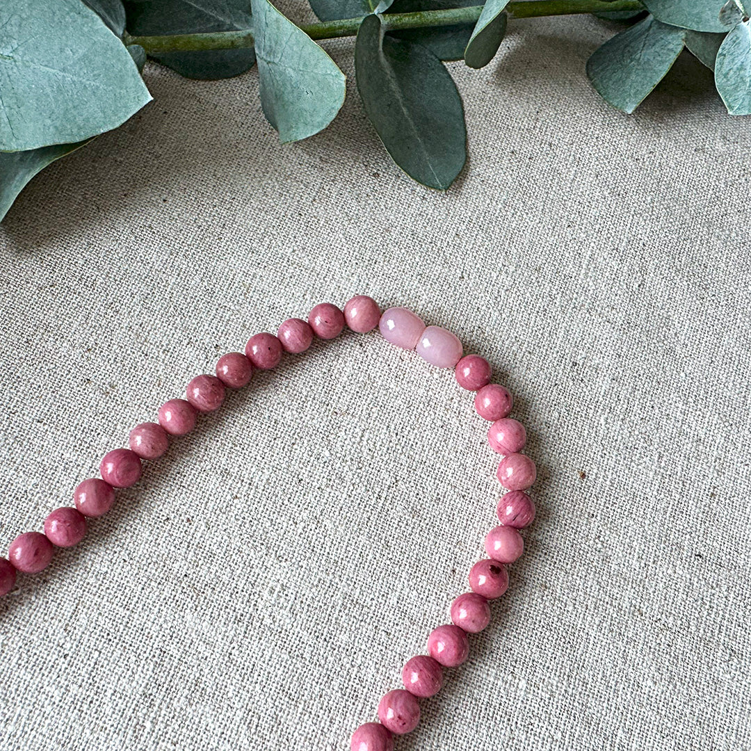 Detail view of the Rhodonite Healing Necklace on natural linen fabric with eucalyptus accents. The image highlights the distinctive lighter pink oval accent beads and the stone's signature rose coloration with subtle grey and black mineral inclusions. Each polished bead displays unique patterning that captures light beautifully across its smooth surface. This healing piece serves as both a striking accessory and a wearable talisman for emotional balance, trauma recovery, and fostering self-worth.