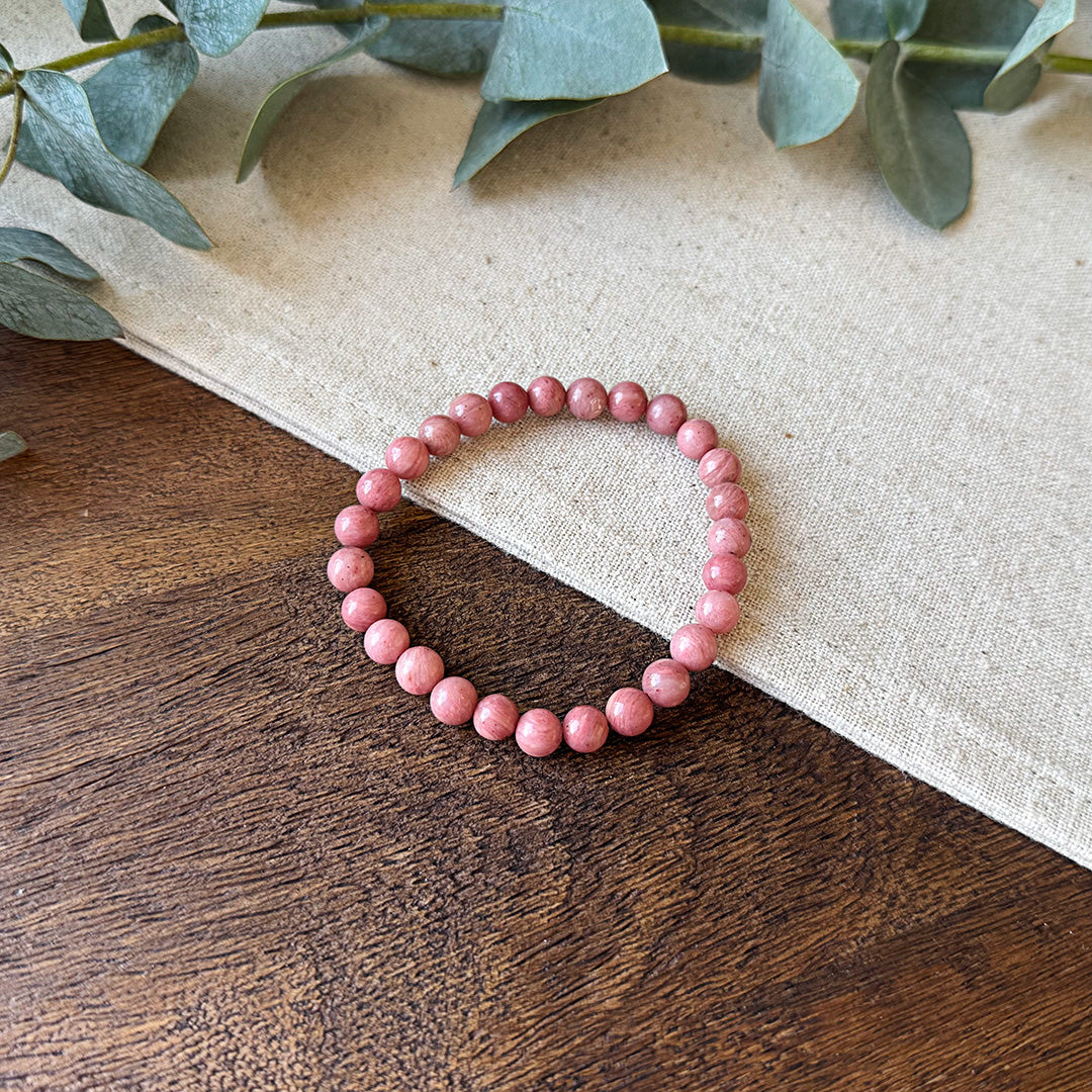 Aventari Rhodonite Wellness Bracelet featuring dusty pink beads resting on a natural wood and linen surface with eucalyptus.