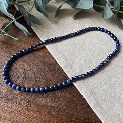 Lapis Lazuli Protection Necklace elegantly arranged on rustic wood and natural linen fabric. The polished gemstone beads showcase a stunning deep royal blue color with characteristic gold pyrite flecks that shimmer in the light. Fresh eucalyptus sprigs frame the upper corner, complementing the necklace's natural beauty while emphasizing its protective and calming qualities. Each bead displays the stone's distinctive rich coloration, creating a striking piece that combines visual appeal with traditional prop