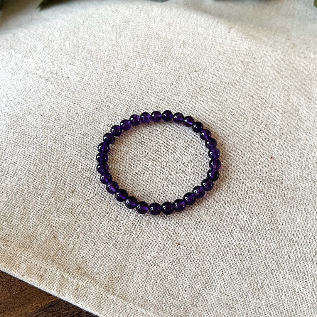 Simple flat lay view of the Aventari Amethyst Bracelet showing its smooth purple beads in a circle on a textured linen background.