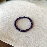 Simple flat lay view of the Aventari Amethyst Bracelet showing its smooth purple beads in a circle on a textured linen background.