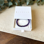 Aventari Amethyst Bracelet presented in its white branded gift box on a light wood and linen surface with eucalyptus accents.