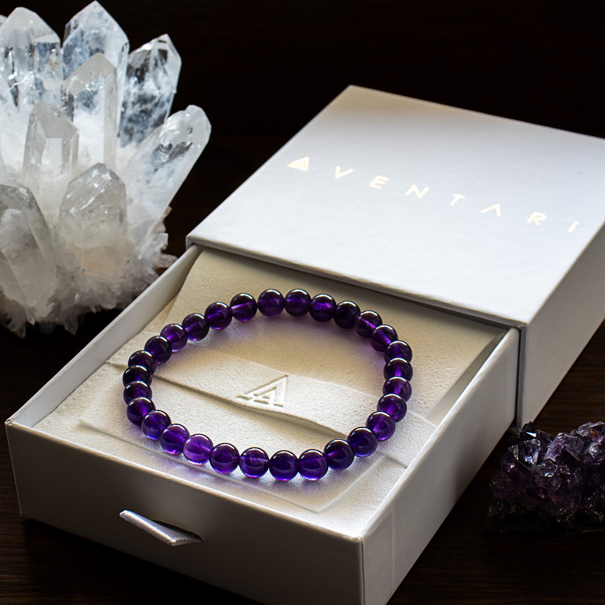 Aventari Amethyst Spiritual Power Bracelet with deep purple beads in its white gift box, displayed next to raw amethyst and clear quartz crystal clusters.