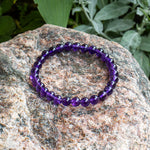 The Aventari Amethyst Spiritual Power Bracelet with its purple beads resting naturally on a textured granite rock outdoors.